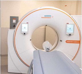 A PET scan machine (it is white and donut shaped with an orange ring around it) with a bench on the end of it in a white room. 